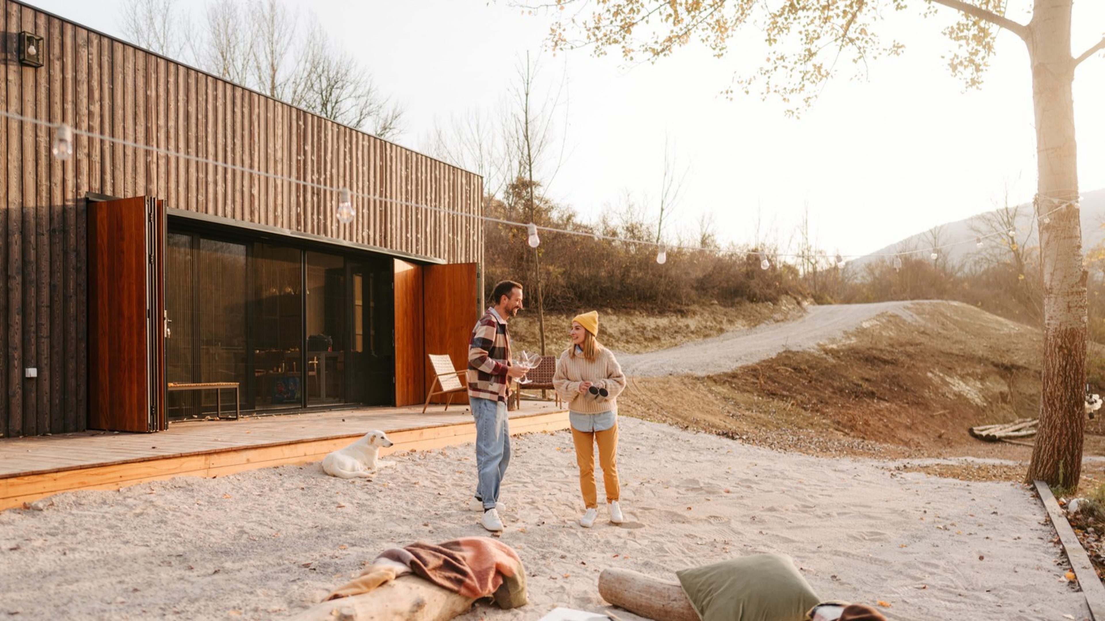 Ein glückliches Paar steht im Garten vor einem modernen Holzhaus und unterhält sich. Es ist in Freizeitkleidung und lacht. Ein Hund liegt im Sand hinter ihnen, und im Hintergrund sind Bäume und eine Hügelkette zu sehen. Es ist ein sonniger Tag, und das Bild strahlt eine entspannte, glückliche Atmosphäre aus.