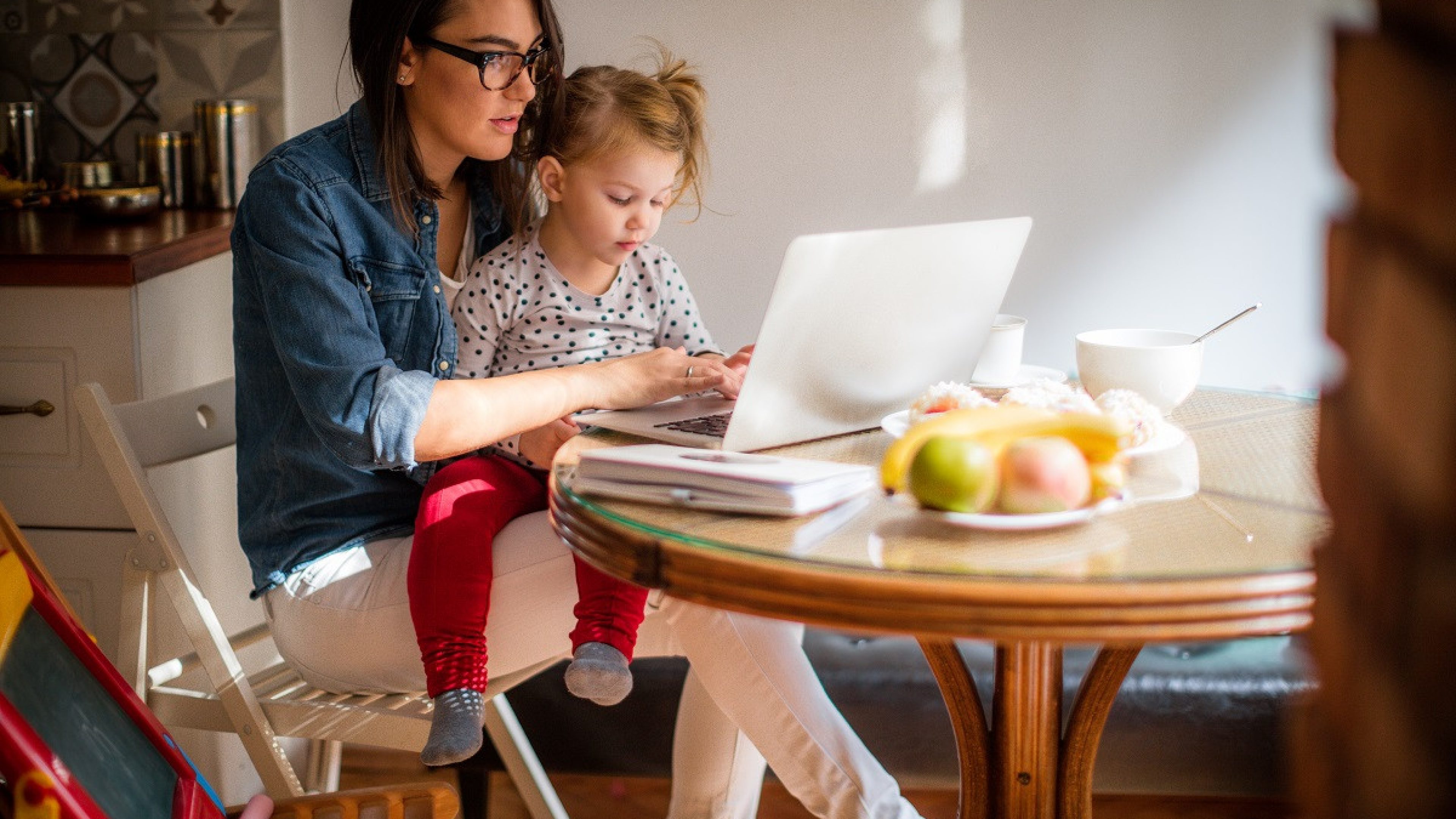 Photo of a little girl trying to help mom who is working on a computer at their dining table