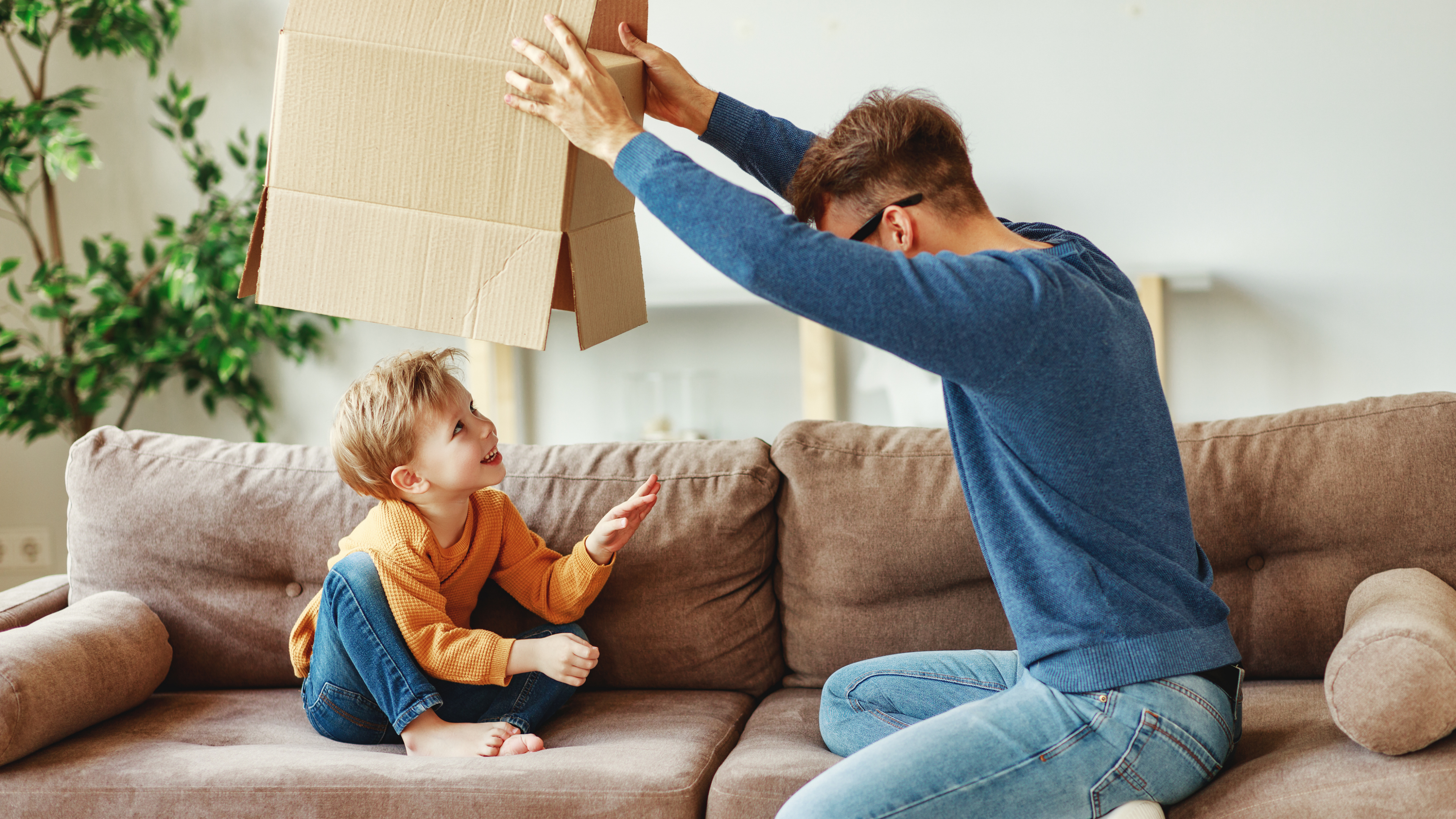 Side view of adult man lifting carton box over smiling boy while sitting on sofa and playing at home together
