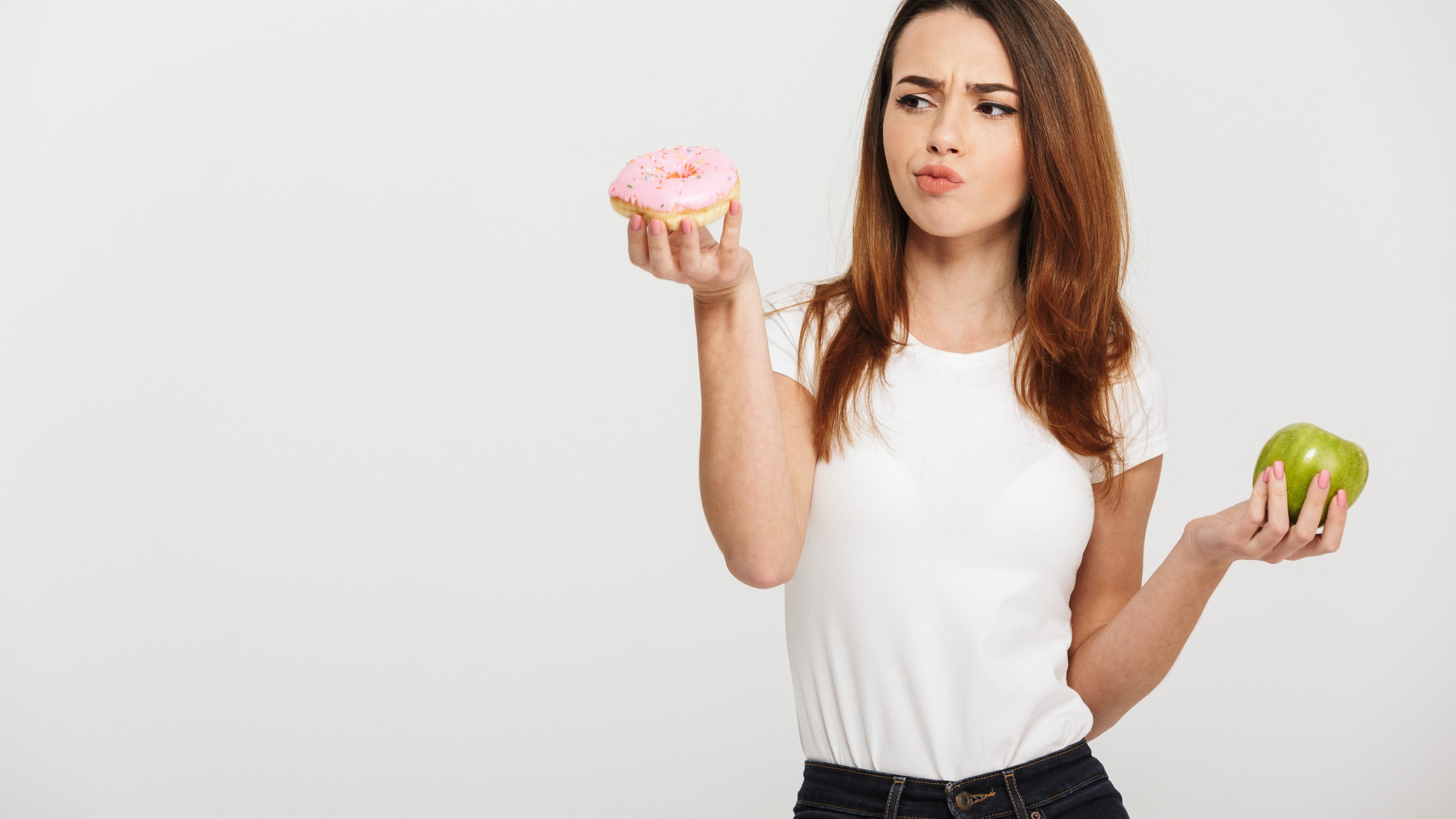Portrait of a confused young woman choosing between donut and green apple isolated over white background