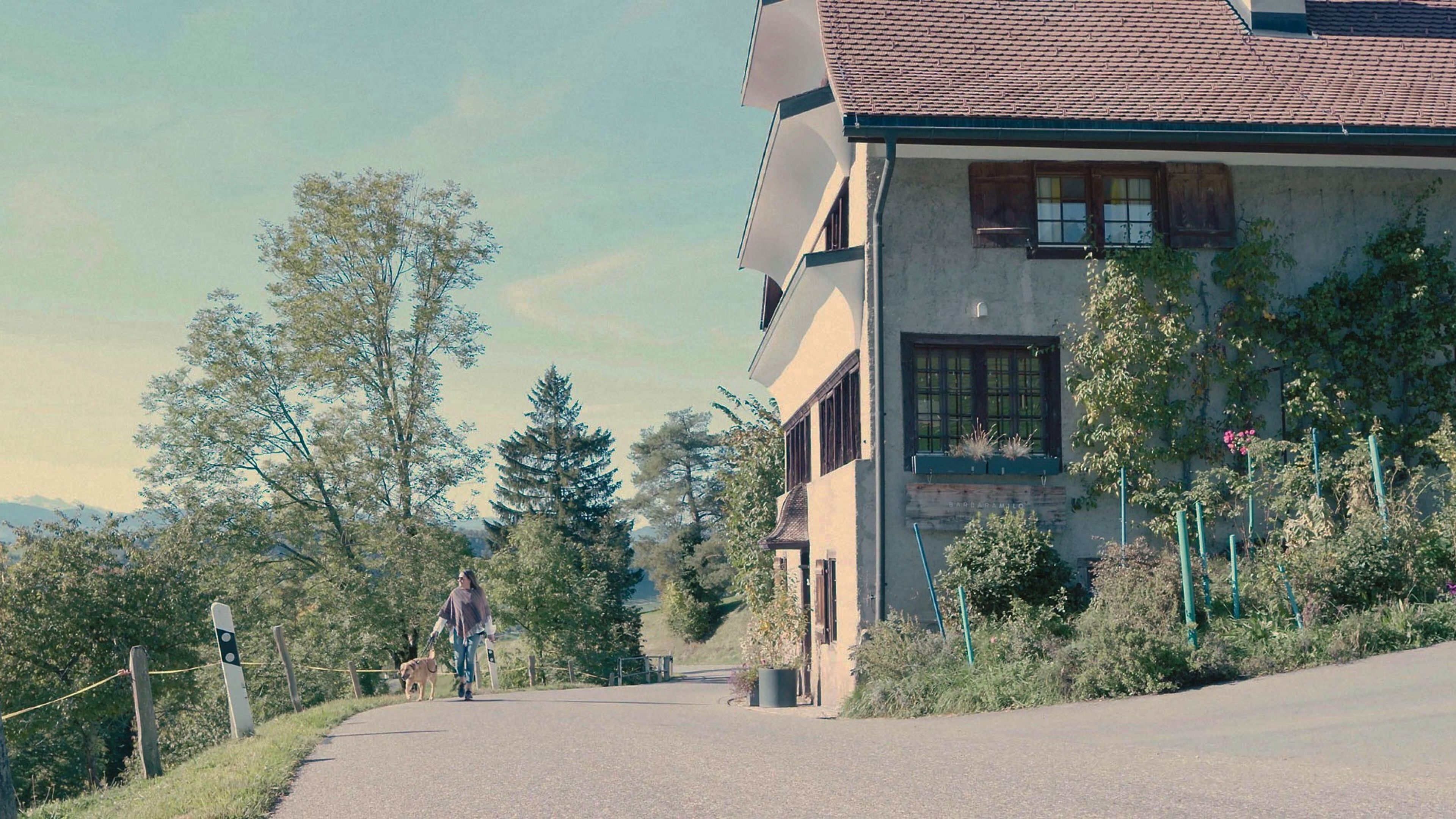 Frau läuft mit Hund an einem Haus vorbei.