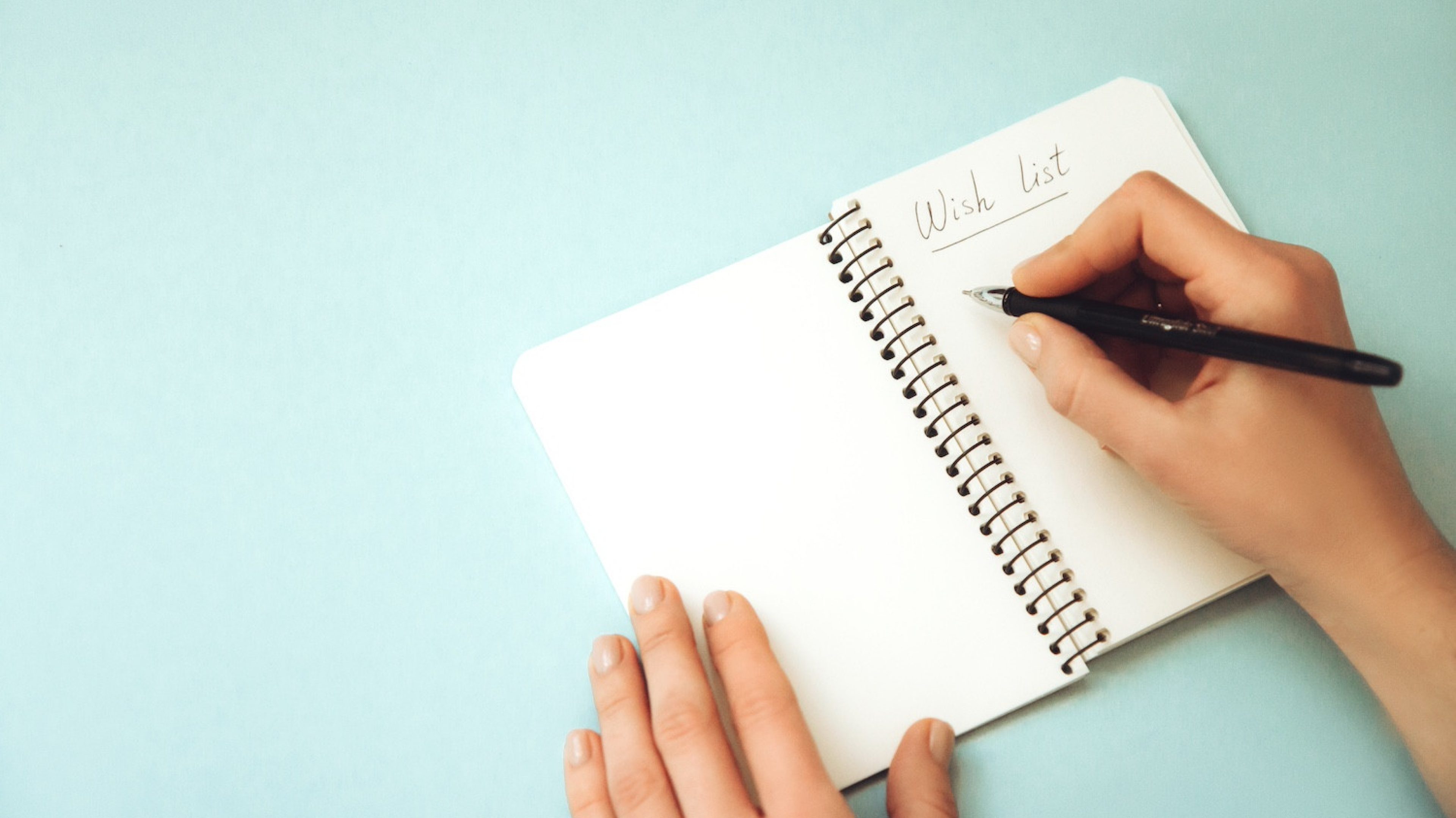 Woman's hand holding pen and open spiral notebook with blank white pages on light background. Wish list