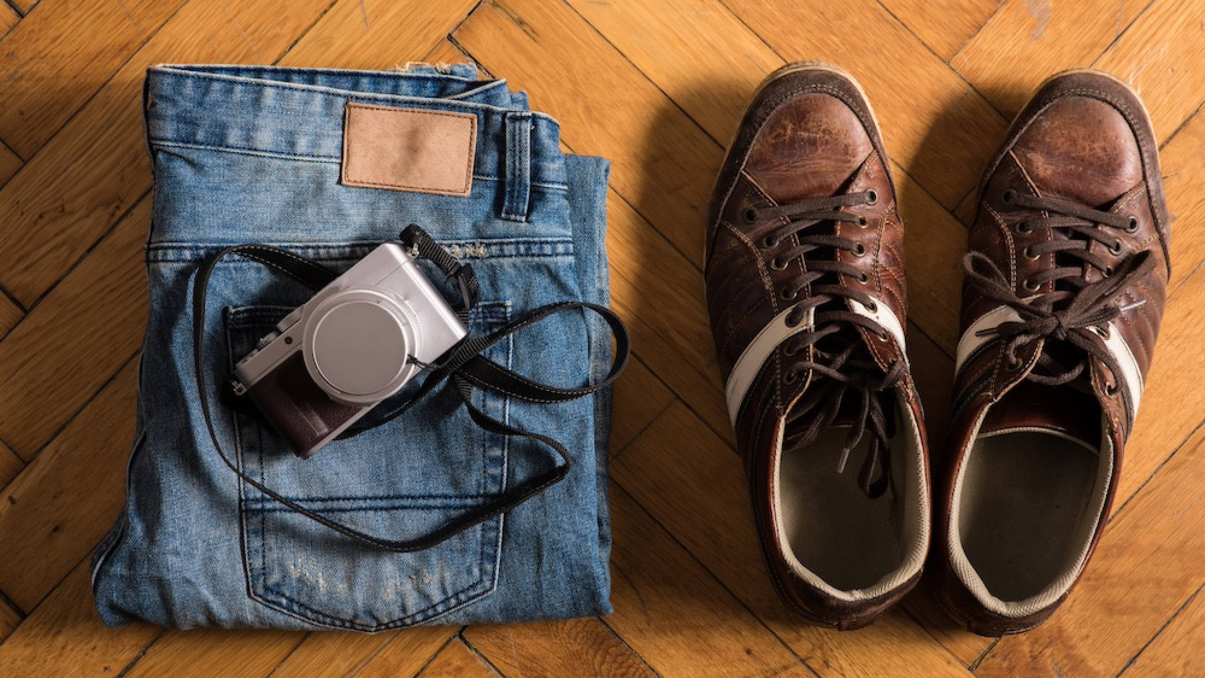 A pair of fretted blue jeans, brown shoes, and a camera lie by on the floor. 