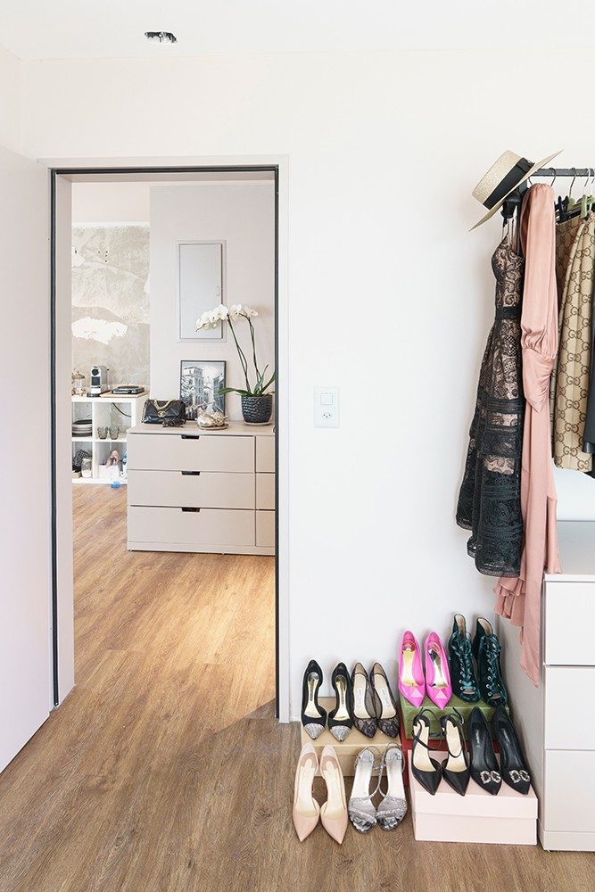 Eine Tür, hinter der sich eine Kommode verbirgt, neben der Tür stehen Schuhe aufgereiht auf Schuhschachteln und eine Kleiderstande mit Kleidern daran.  