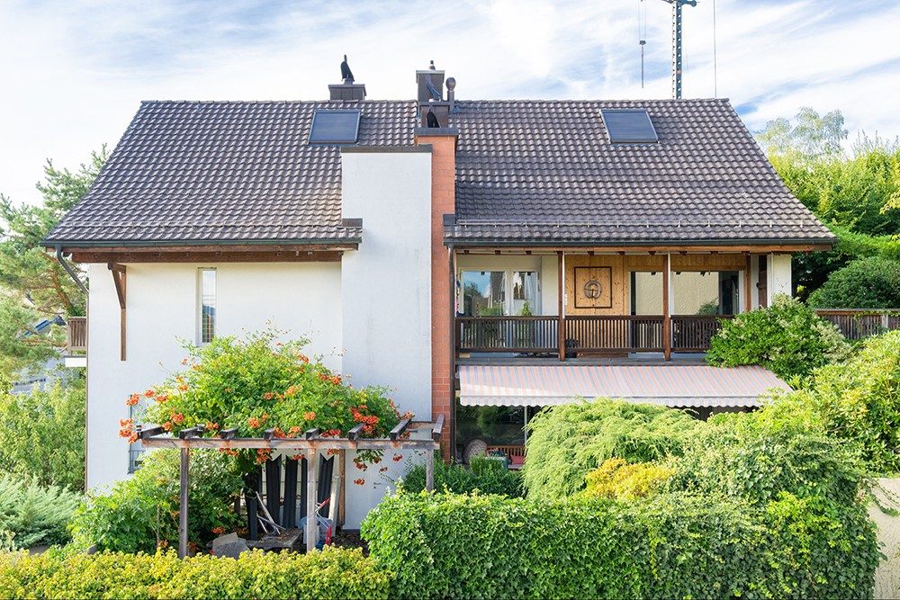 Ein grosser Baum steht vor dem Haus, das Haus ist weiss mit einigen Holzelementen