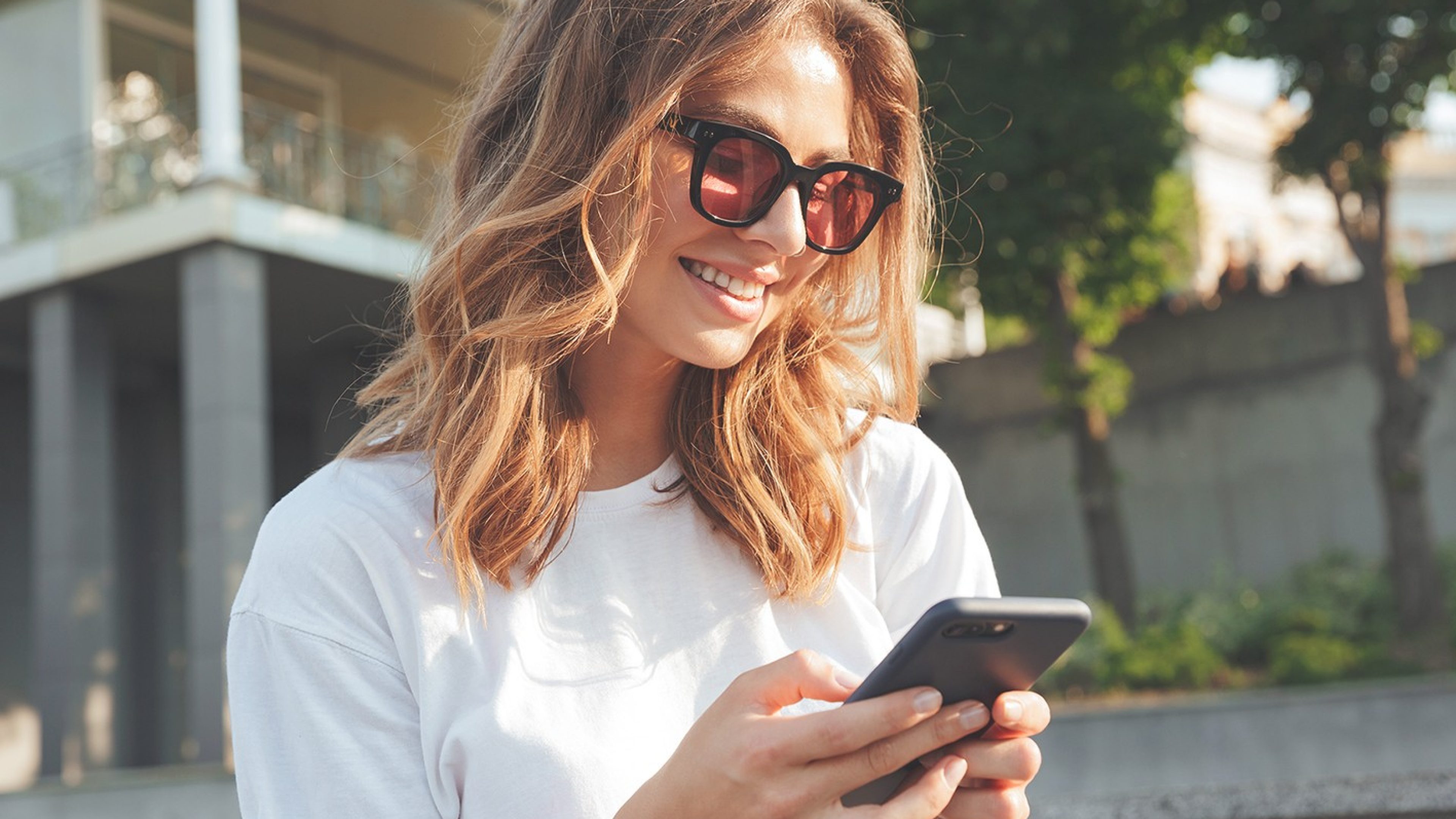Junge Frau mit Sonnenbrille blickt auf ihr Smartphone und lächelt
