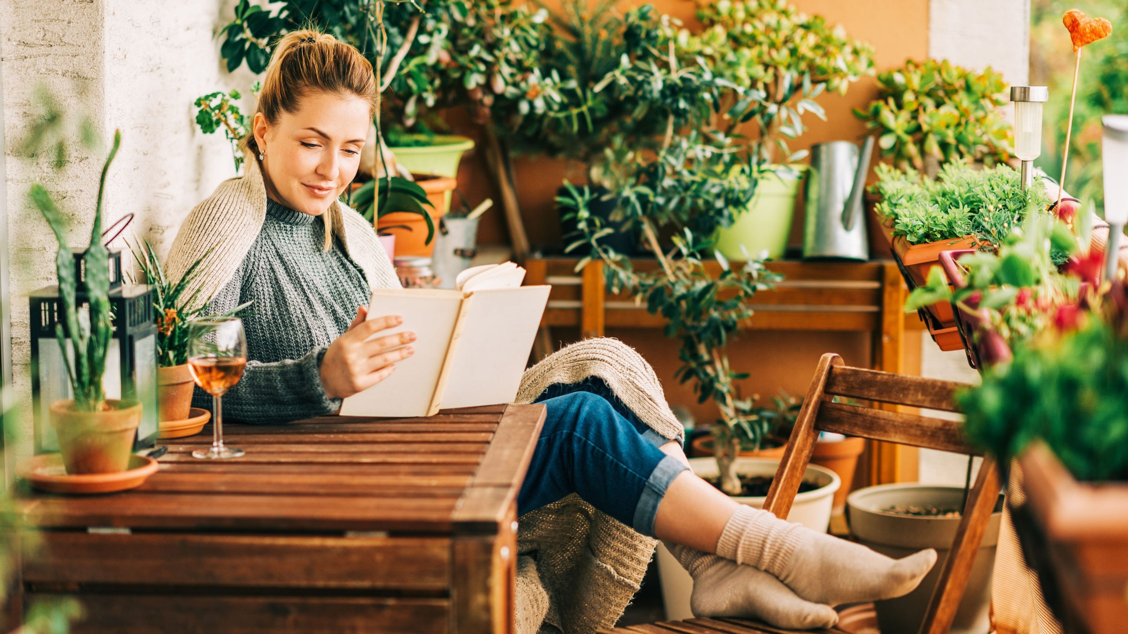 Young beautiful woman relaxing on cozy balcony, reading a book, wearing warm knitted pullover, glass of wine on wooden table