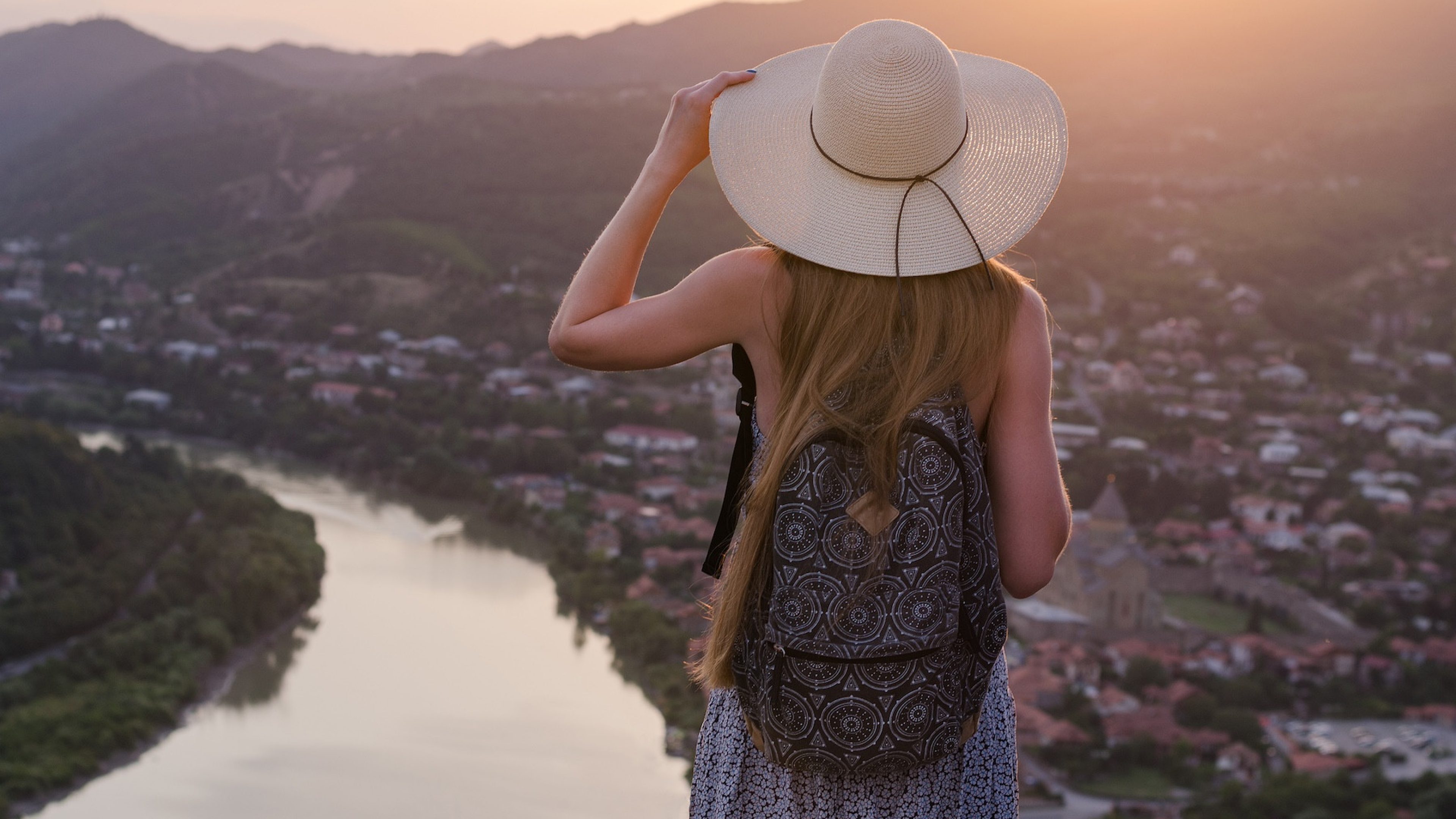 Young beautiful girl in dress and hat, looking at river and city sunset