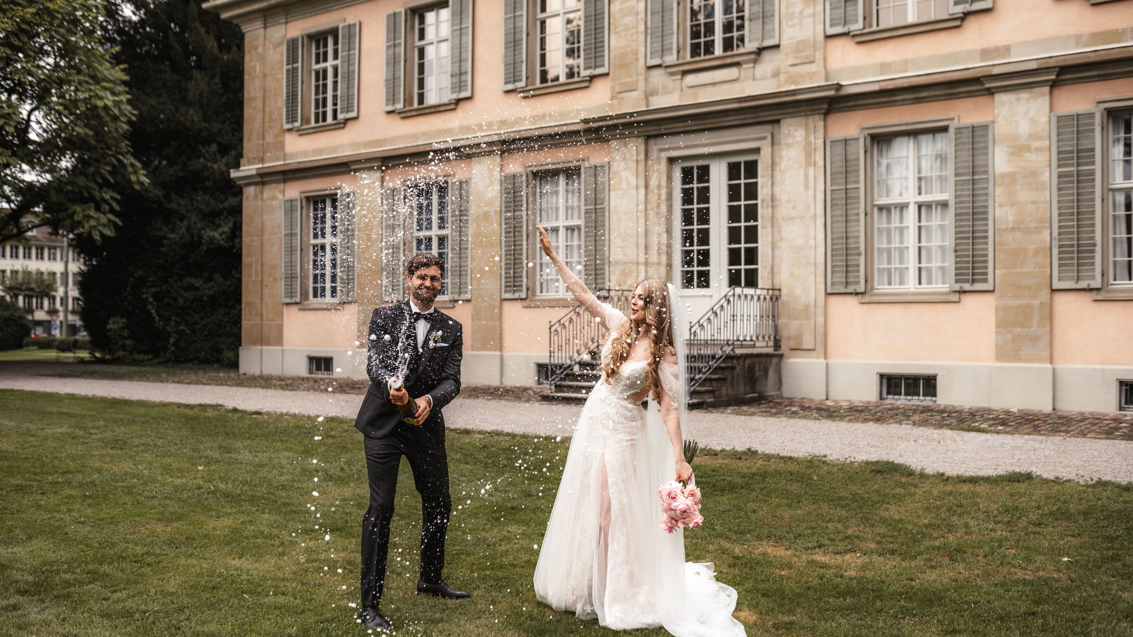 La mariée lève son bouquet de fleurs avec joie, le marié lui fait signe, un grand sourire sur les lèvres. Ils descendent ensemble les escaliers d’un bâtiment historique. Sa posture inspire la joie et la confiance.  La mariée porte une robe en dentelle blanche et a de longs cheveux blonds bouclés coiffés d’un voile blanc.
