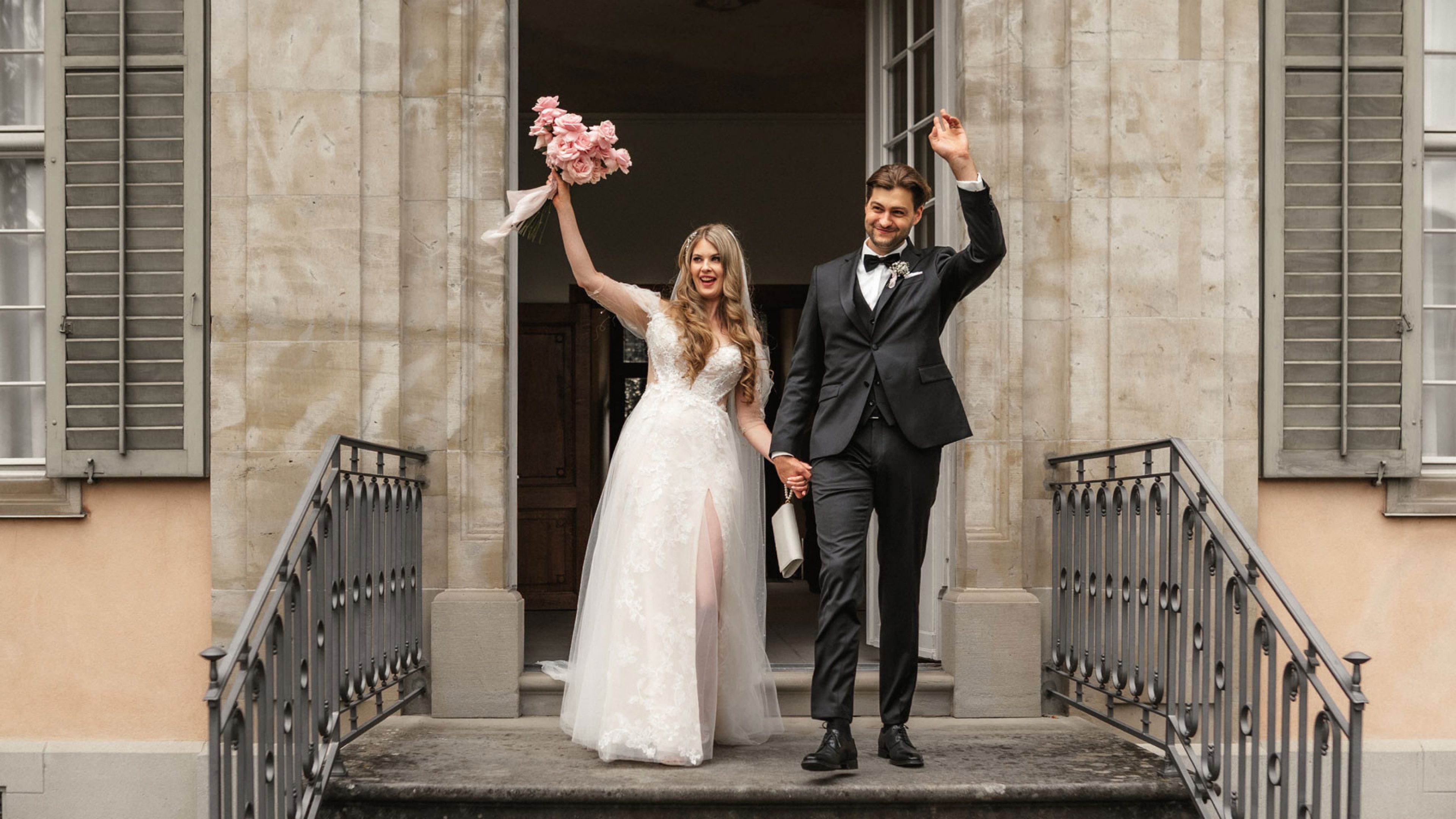 Un couple fraîchement marié quitte l’office de l’état civil par un escalier en pierre. La mariée porte une robe en dentelle blanche et tient un bouquet de roses roses dans la main. Elle a de longs cheveux blonds bouclés couverts d’un voile blanc. Ils se regardent, rayonnants, et se tiennent l’un l’autre alors qu’ils descendent les escaliers. L’architecture du bâtiment en arrière-plan est classique avec des colonnes claires et de grandes fenêtres.