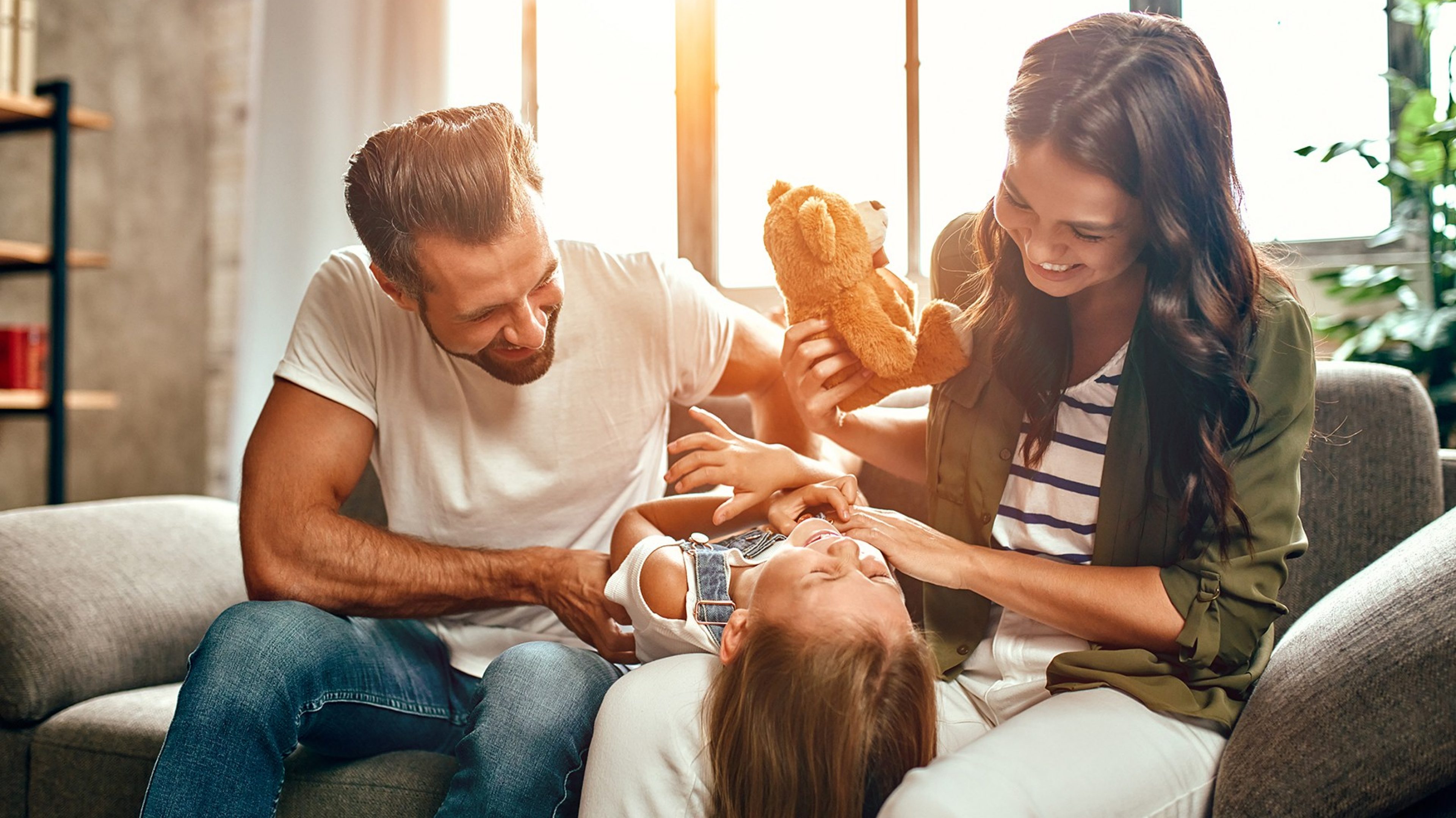 Happy dad and mom with their cute daughter and teddy bear hug, play and have fun sitting on the sofa in the living room at home.