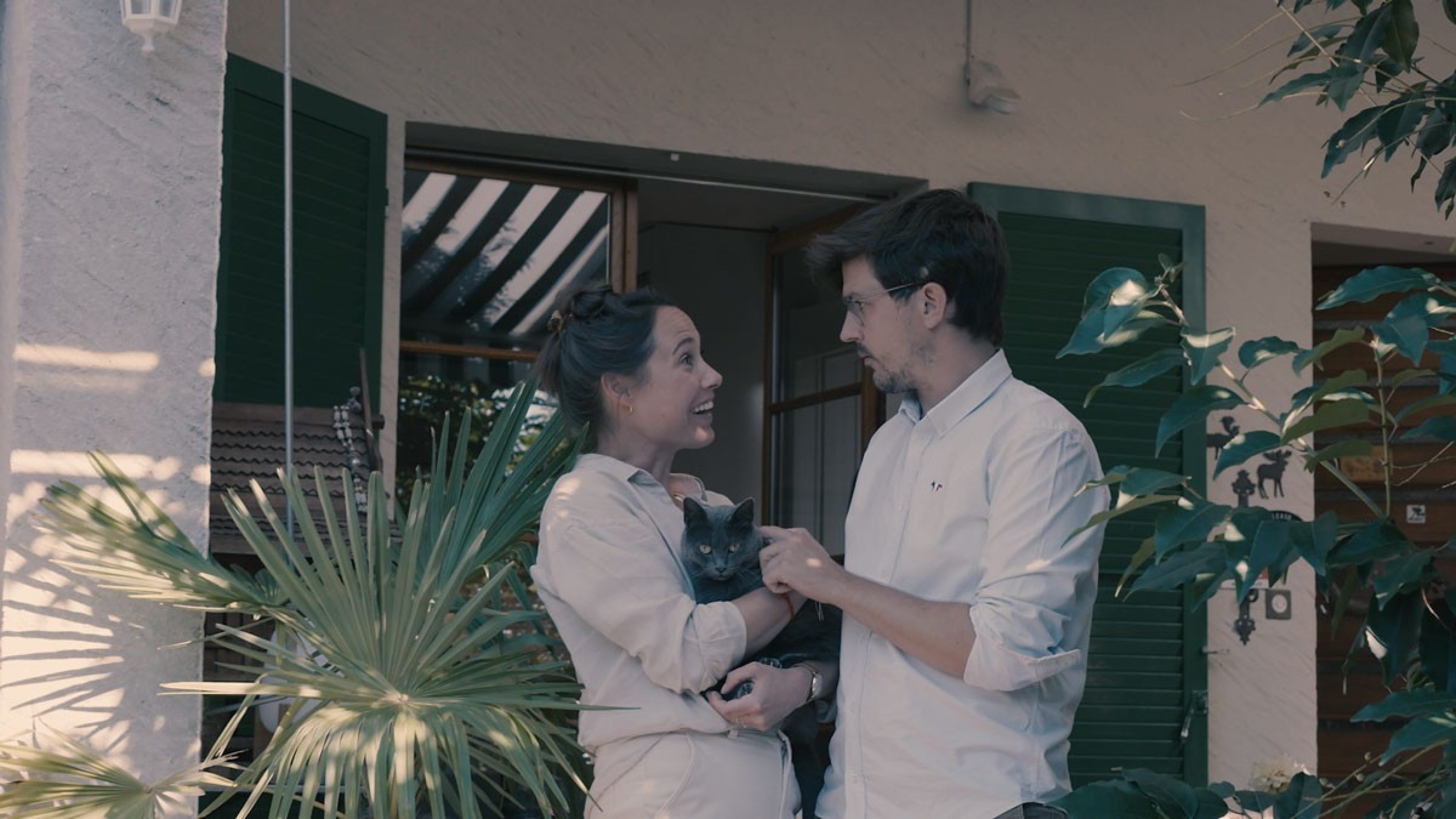 Un couple avec un chat dans les bras devant une maison