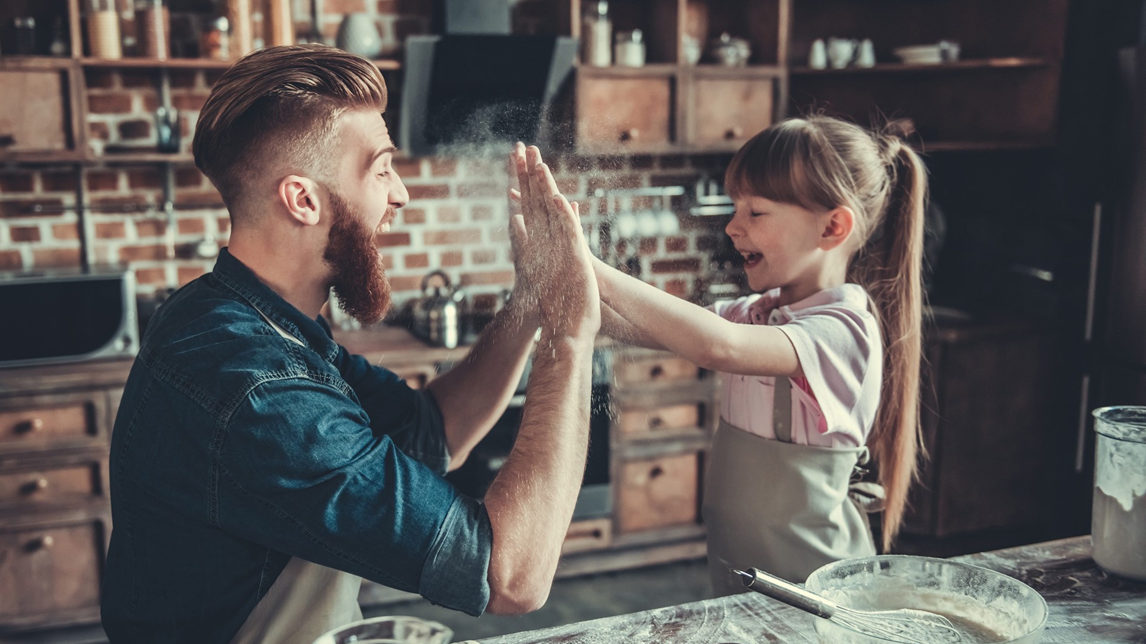 Cute little girl and her handsome bearded dad in aprons are having fun with flour, giving high five and smiling while cooking in kitchen