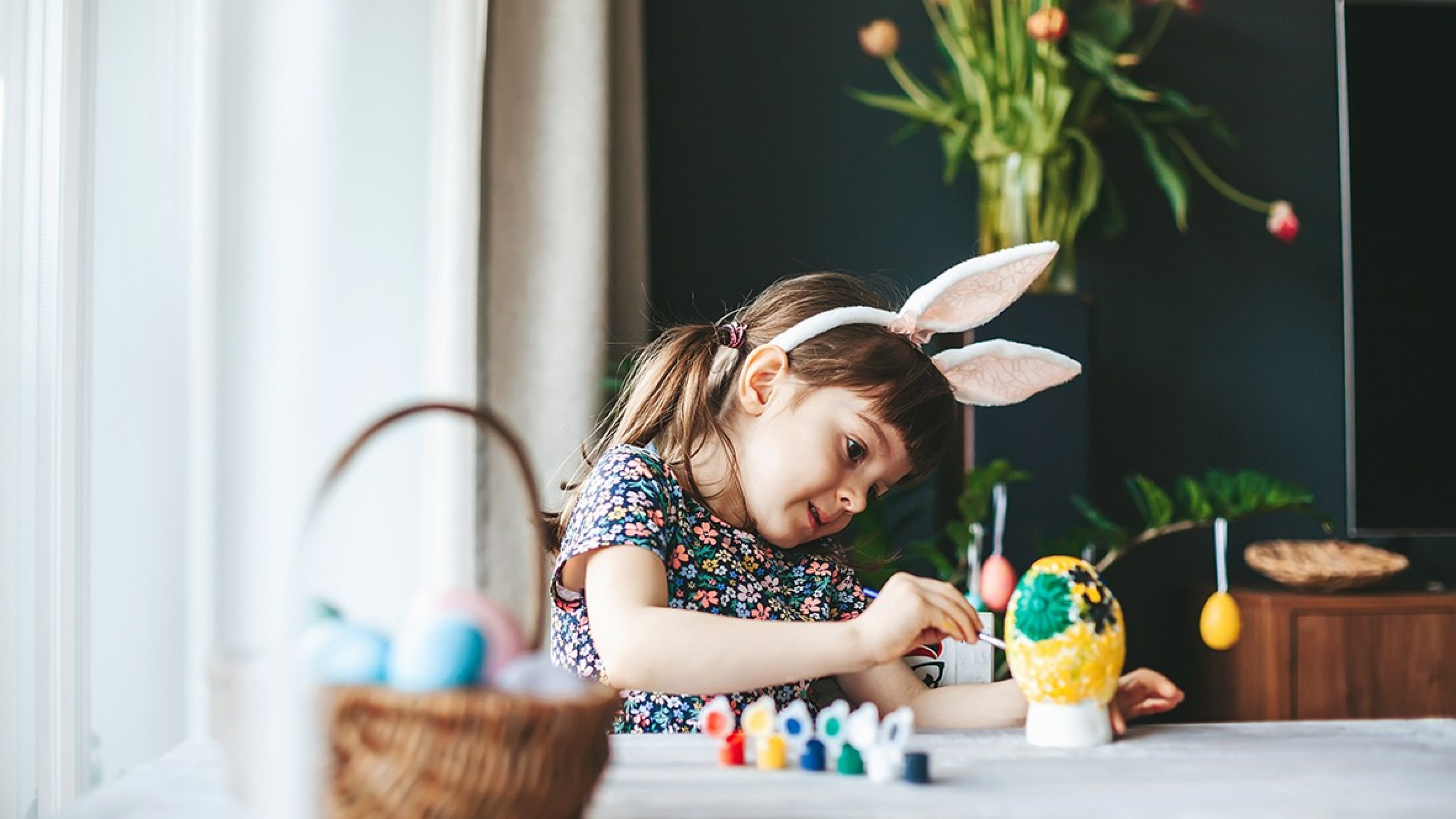Little girl with bunny ears painting gypsum Easter egg with paintbrush. Easter celebration concept.