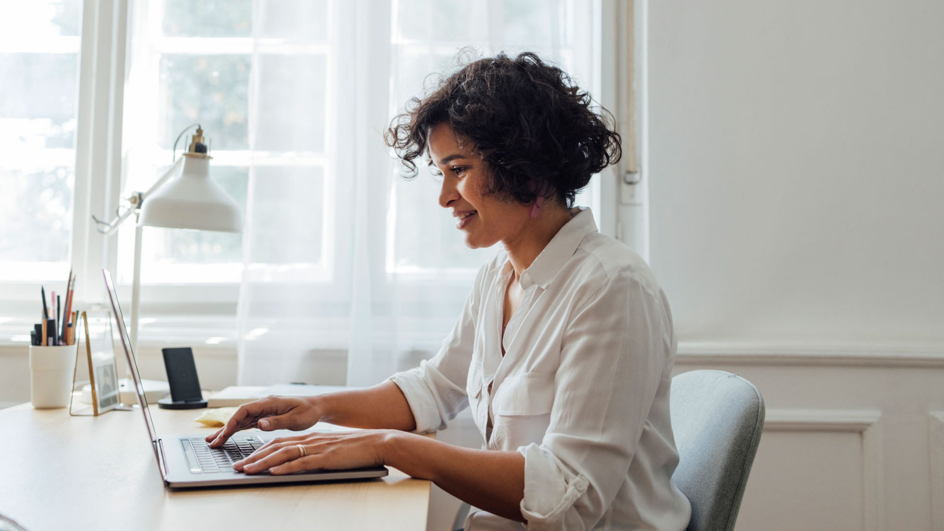 Une jeune femme souriante est assise dans un café moderne et travaille de manière concentrée sur son ordinateur portable. Devant elle, une tasse de café et quelques feuilles blanches sont posées sur la table. La pièce est bien éclairée, dans une atmosphère élégante et avec de grandes fenêtres inondant la pièce de lumière naturelle. 
