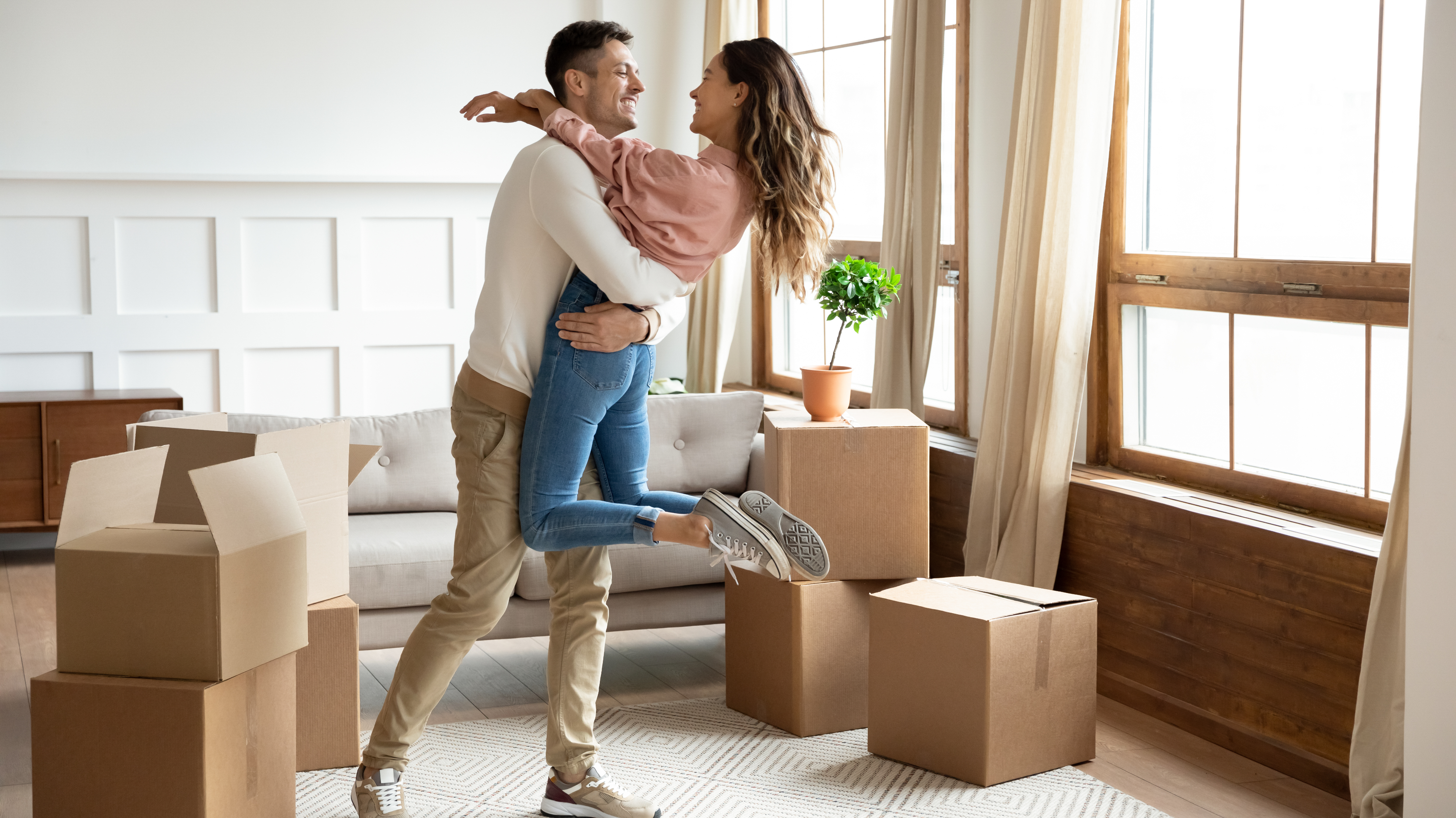 Happy young husband lifting excited wife celebrating moving day with cardboard boxes, proud overjoyed family couple first time home buyers renters owners having fun enjoy relocation, mortgage concept