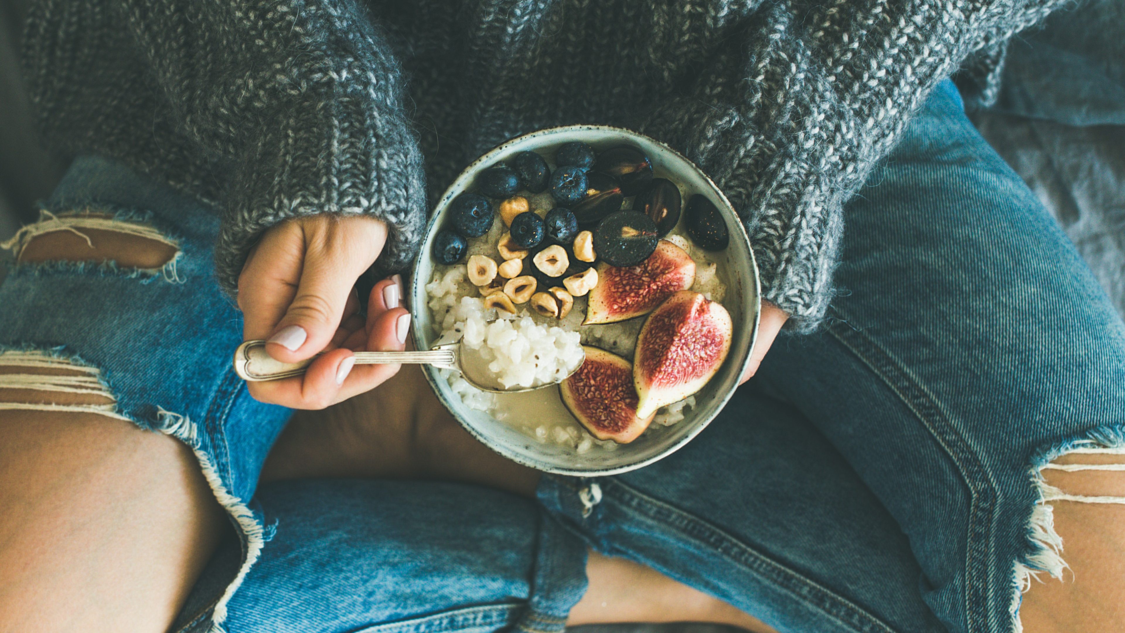 Healthy winter breakfast in bed. Woman in woolen sweater and shabby jeans eating rice coconut porridge with figs, berries, hazelnuts, top view. Clean eating, vegetarian, comfort food concept