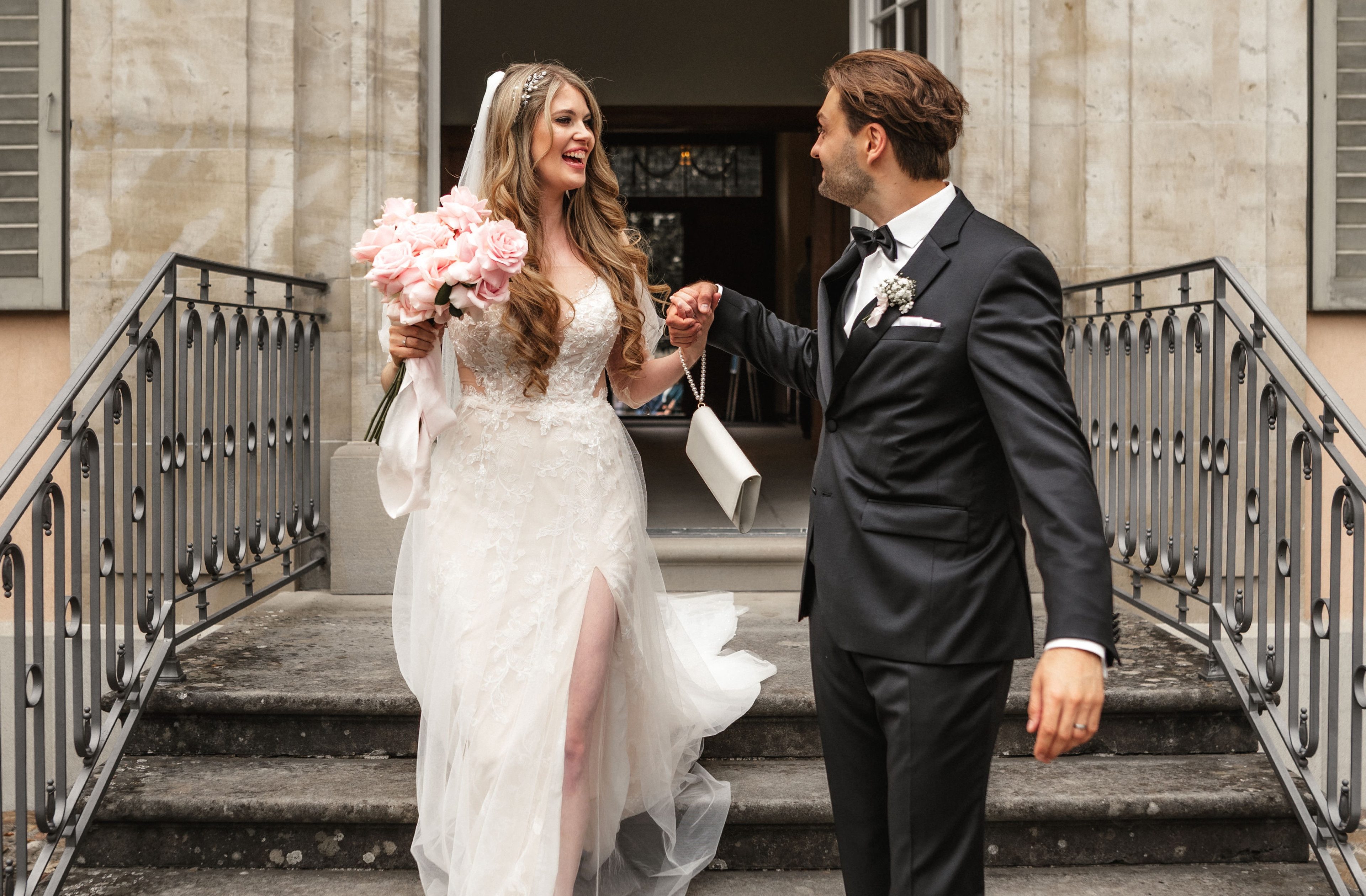 Die Braut steht im Vordergrund eines Pavillons, leicht zur Seite gewendet, mit verträumtem Blick. Sie trägt ein elegantes Kleid mit Tüllärmeln und eine Haarverzierung. Im Hintergrund steht der Bräutigam in Anzug und Fliege, die Hände vor dem Körper gefaltet.