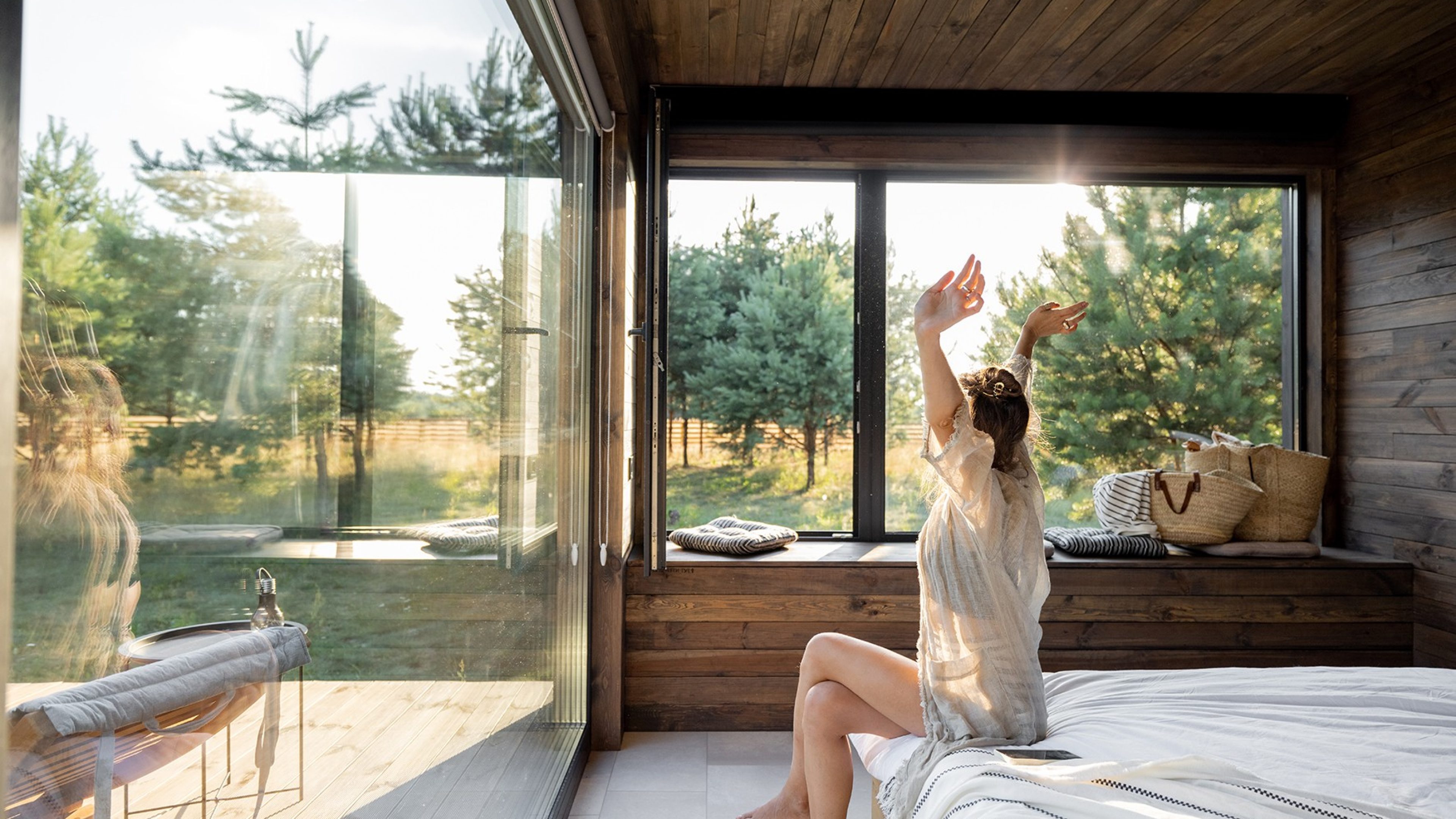 Woman wakes up in a country house or hotel with panoramic windows in pine forest raised her hands yawning. Good morning and recreation on nature concept