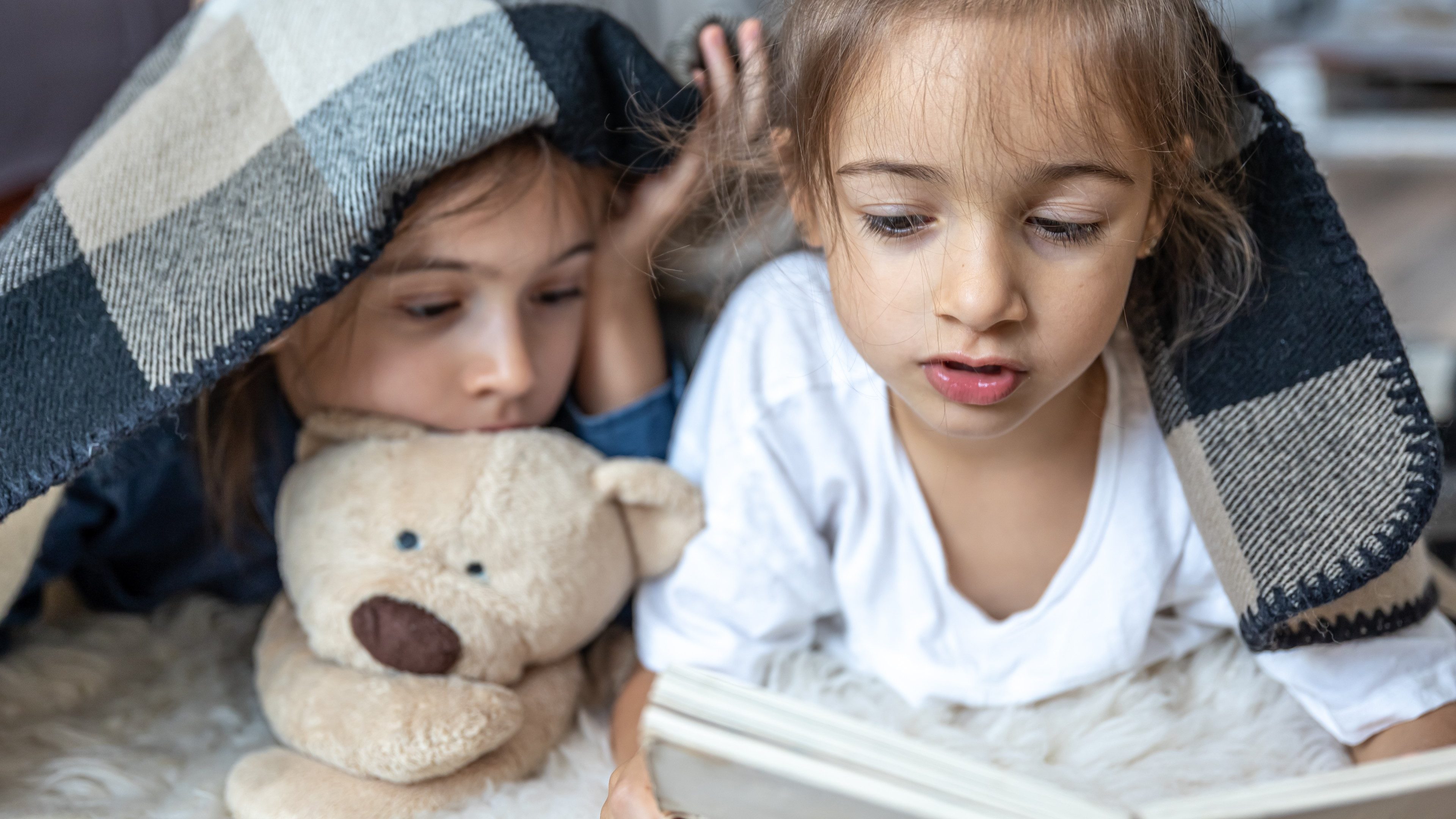 Little sisters are reading a book with a teddy bear lying on the floor in the room.