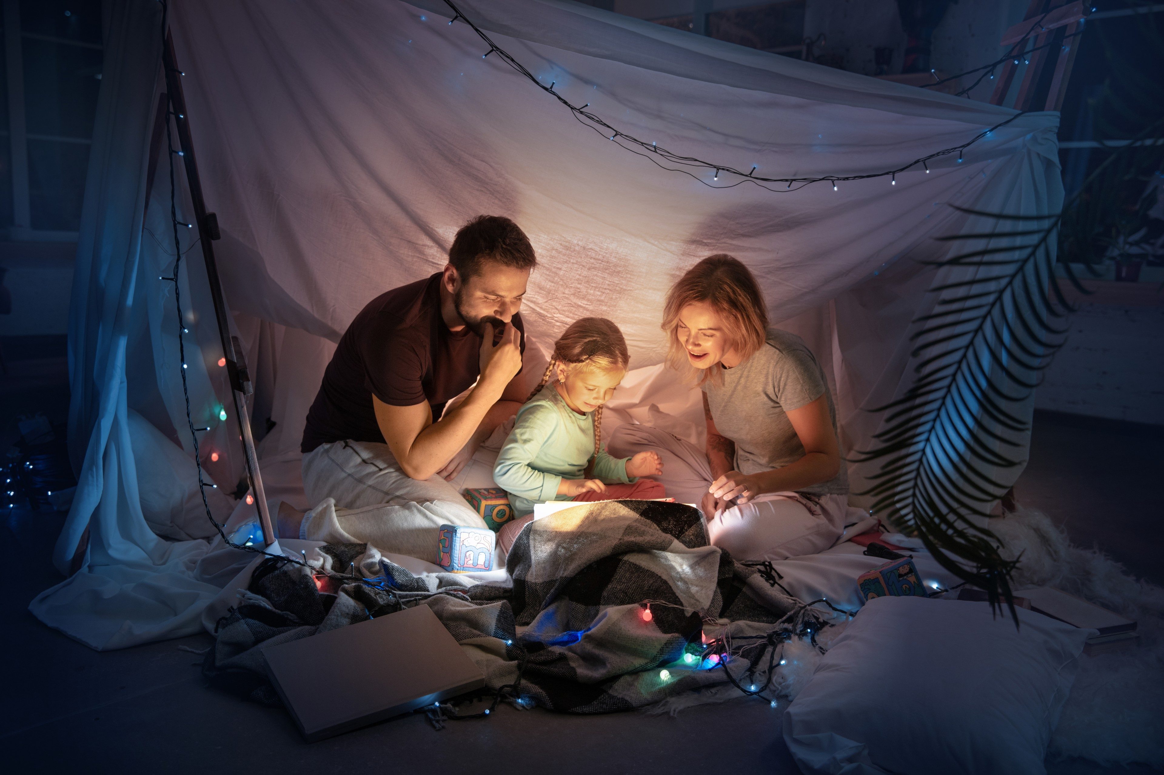 Caucasian family sitting in a teepee, reading stories with the flashlight in dark room with toys and pillows. Caucasian models. Home comfort, family, love, Christmas holidays, storytelling time.