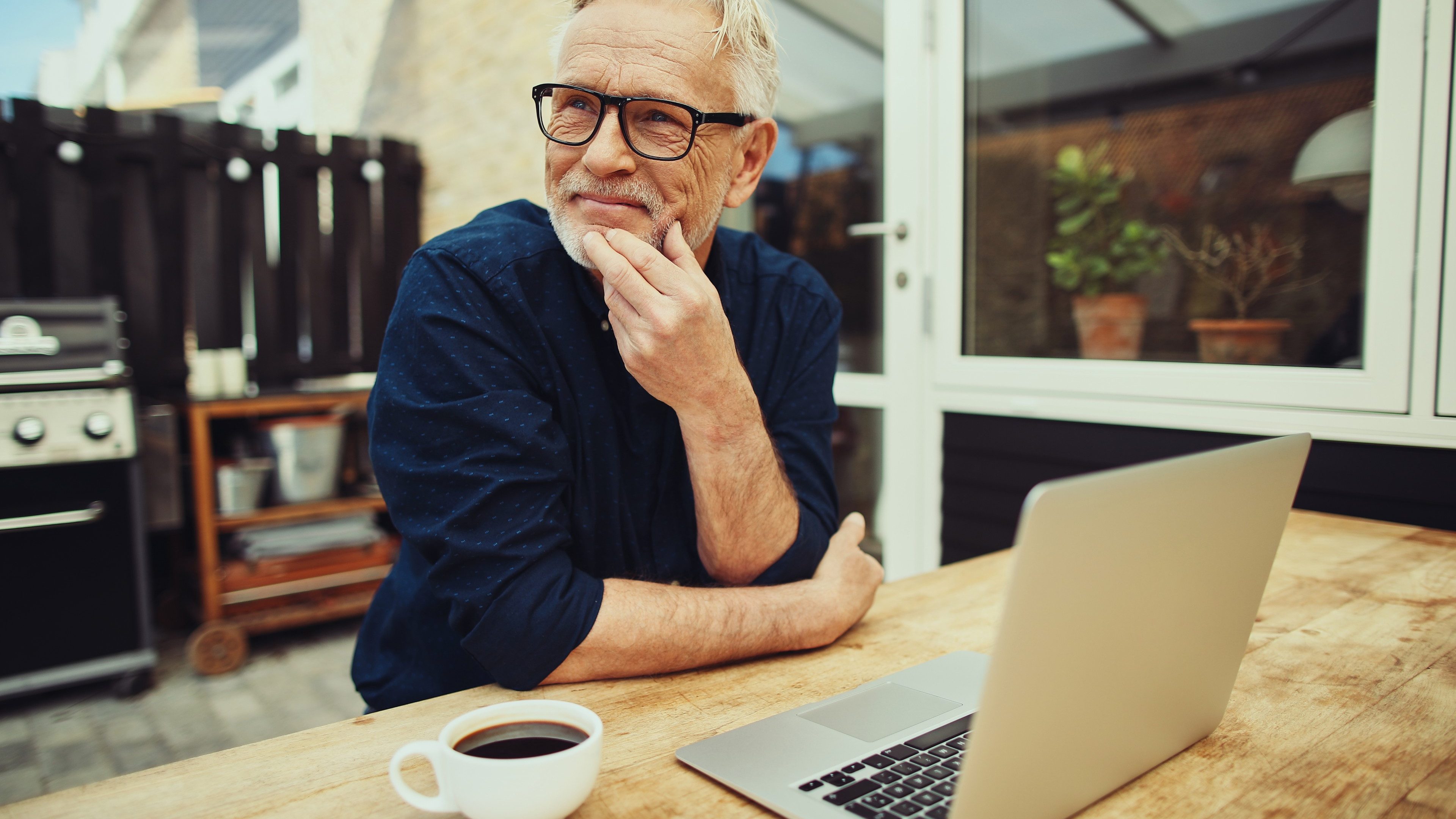 Smiling senior man sitting at a table outside on his patio working online with a laptop and drinking a cup of coffee