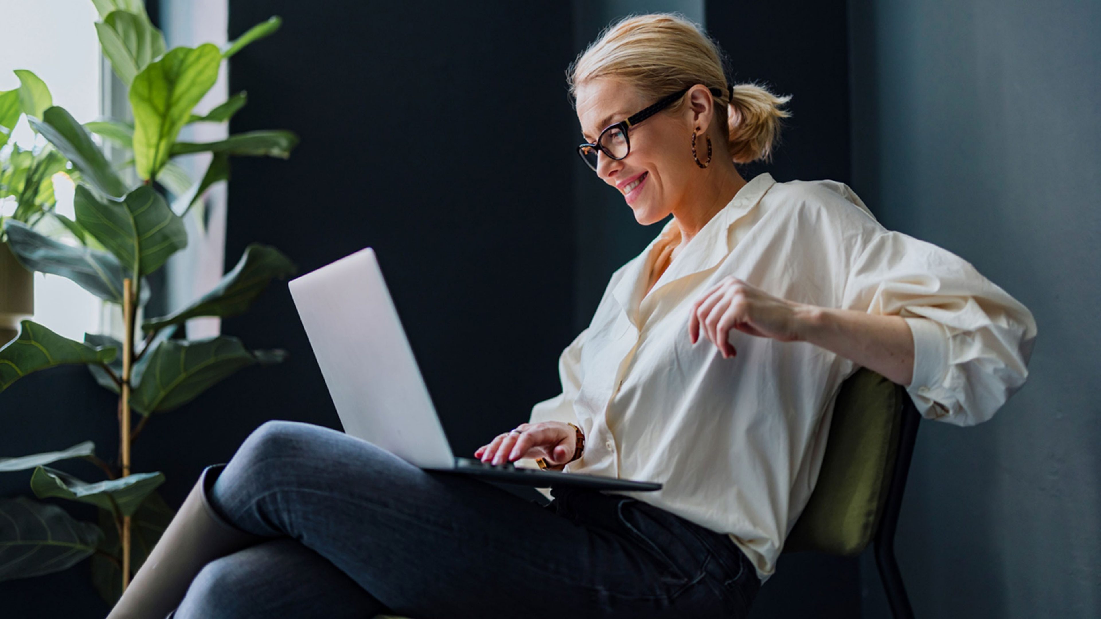 Una donna con occhiali e orecchini è seduta su una poltrona con un PC portatile
