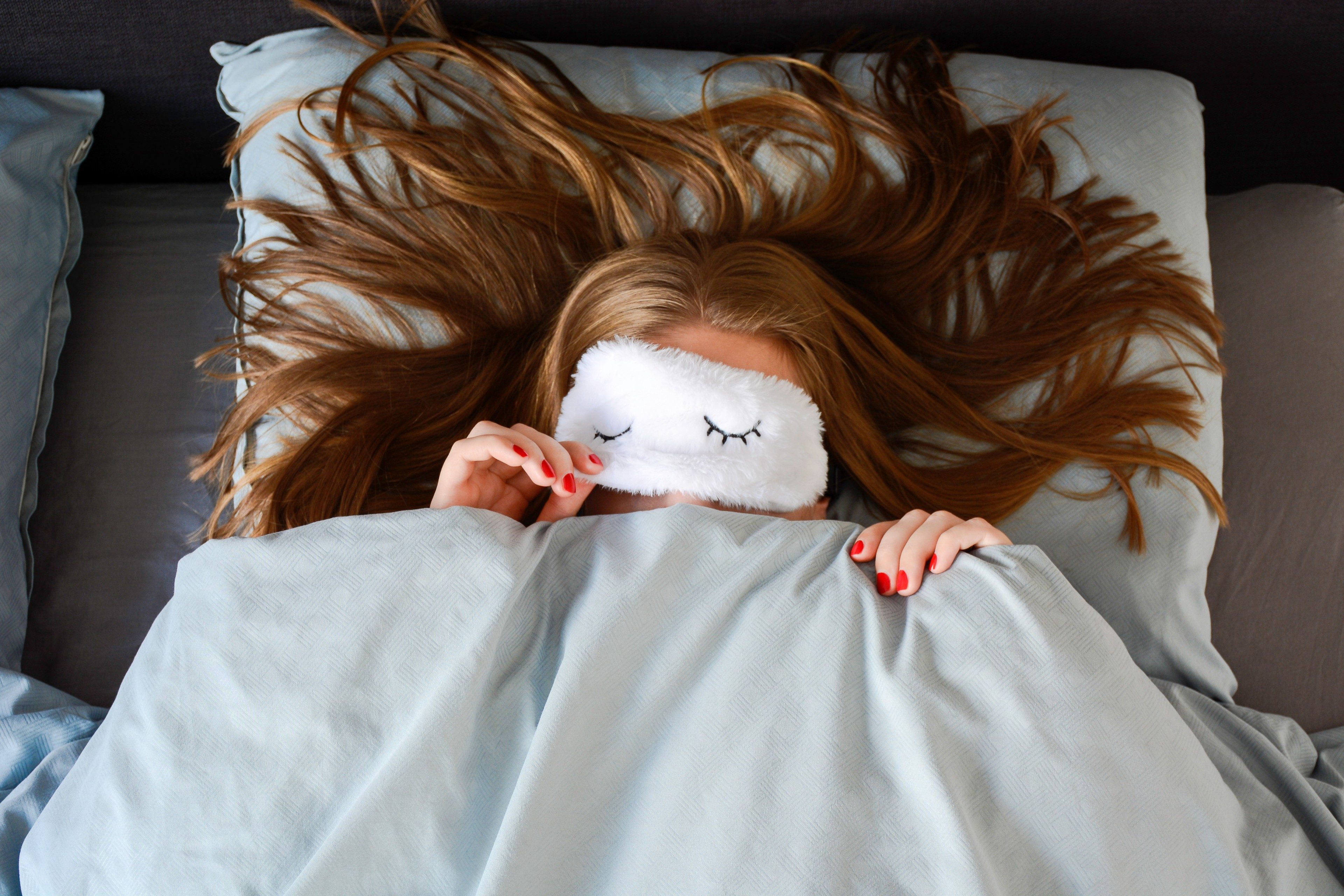 Young woman with long hair and a red manicure is sleeping in bed with sleep mask and peeking from underneath the sleeping mask